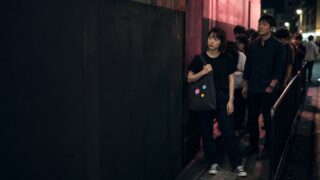 ライブ終演後の物販列で無銘トートを抱く24歳女性と静かに待つ30代男性