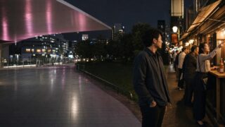 駅前歩道橋から夜の公園越しに暖かな横丁へ向かう30代男性